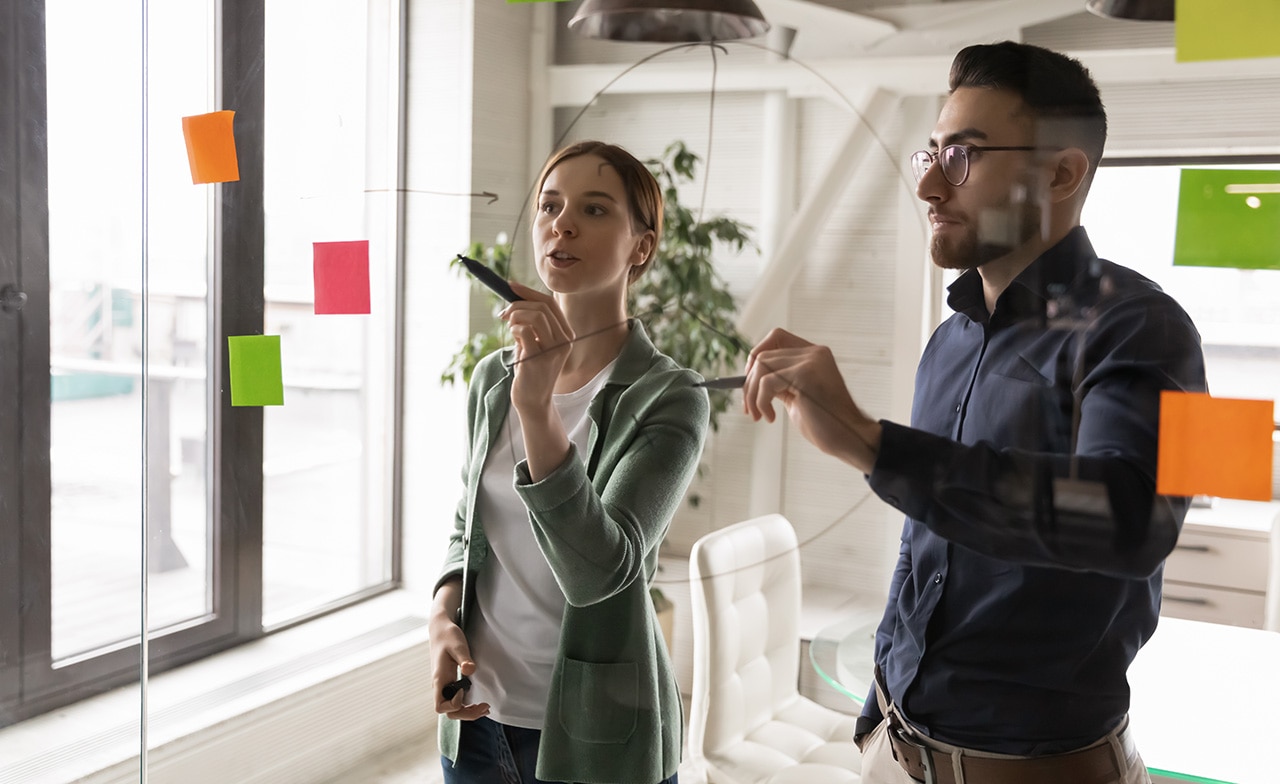 Two professionals writing on clear white board with post-it notes