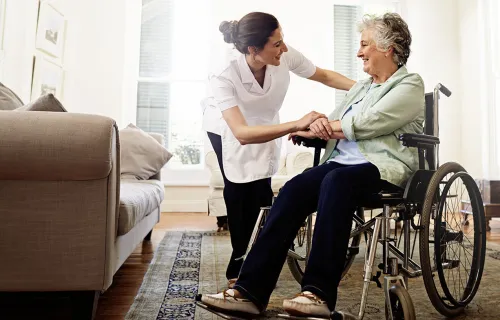 En vårdgivare hjälper en äldre kvinna i rullstol i hemmet, båda ler och har en varm kontakt.