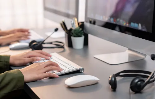 Närbild på händer som skriver på ett tangentbord framför en stationär dator, med headset och…