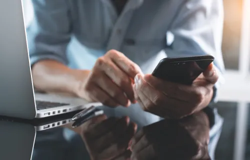 Person in front of a laptop looking at mobile phone.